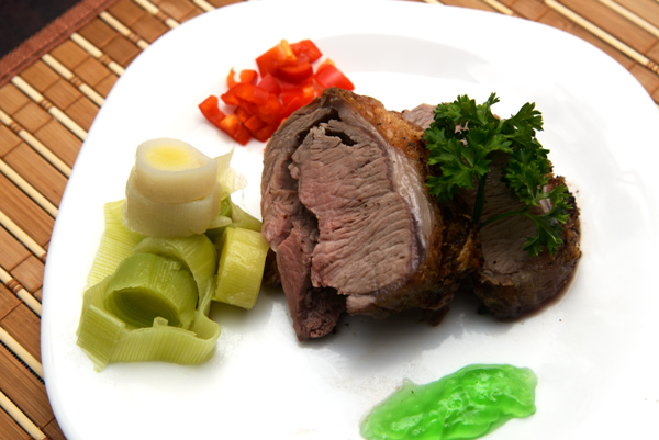 slices of a boneless lamb loin on a plate with steamed leeks, mint sauce and red bell pepper